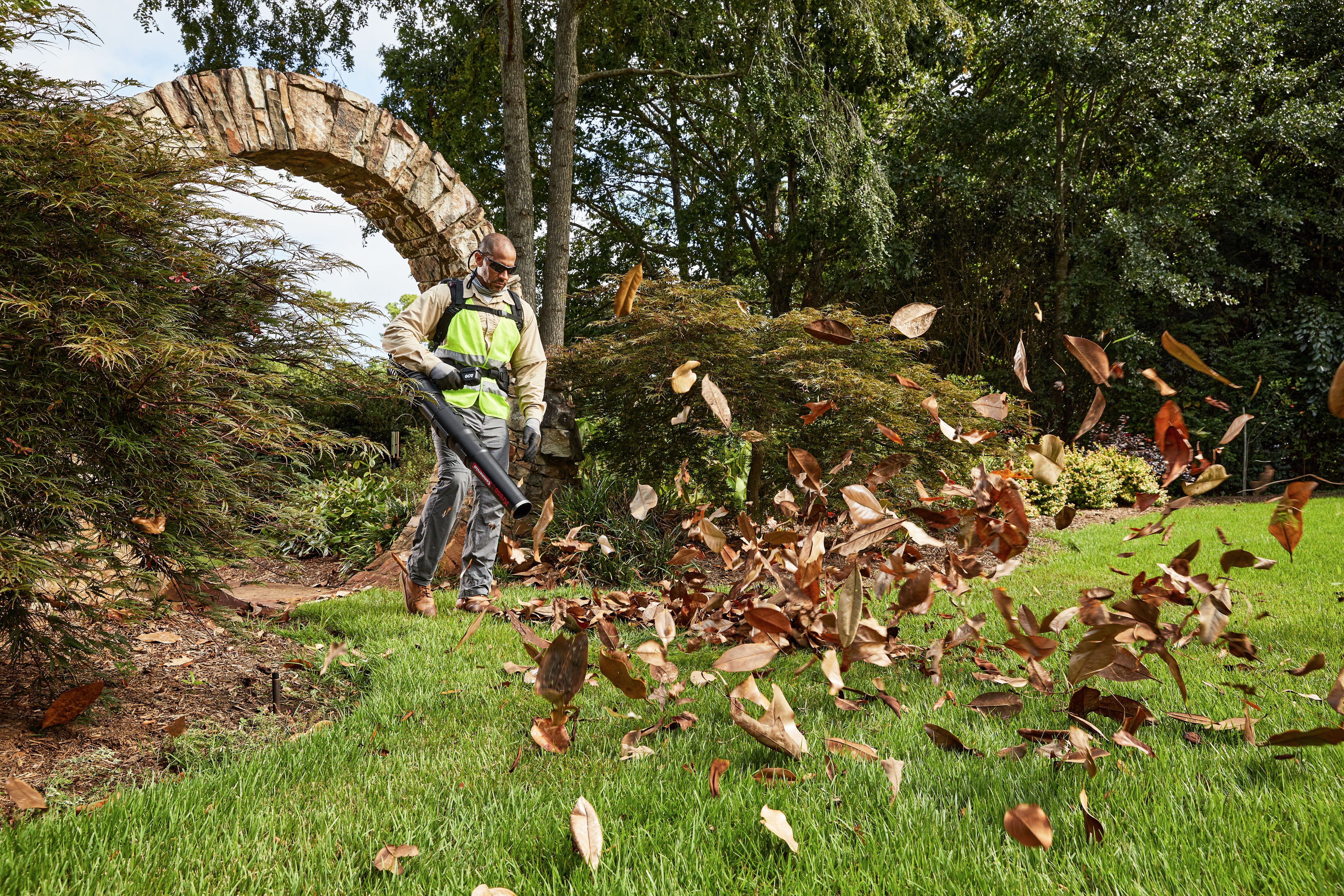 Person using a leaf blower in a garden with trees and an archway in the background