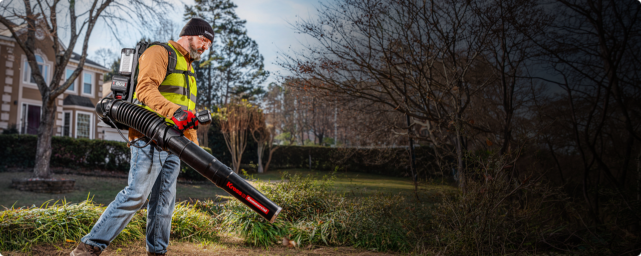 A man wearing a high-visibility safety vest and face mask uses a large black leaf blower with a backpack and red accent nozzle to clear grass and brush in a residential yard.